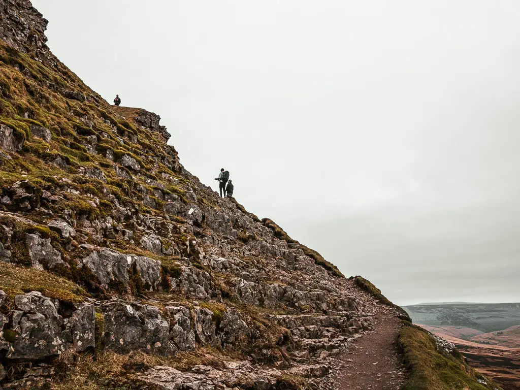 A small dirt trail on the right, along the side of the hill, and lots of rocks up the hill on the left. There are a few people walking up the rocks ahead.