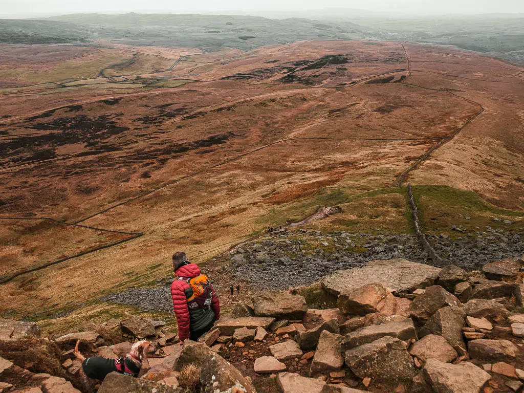 Looking down the rocks, and to the view below, on the walk up to the top of Pen-y-Ghent. There is a man in a red jacket looking down. 