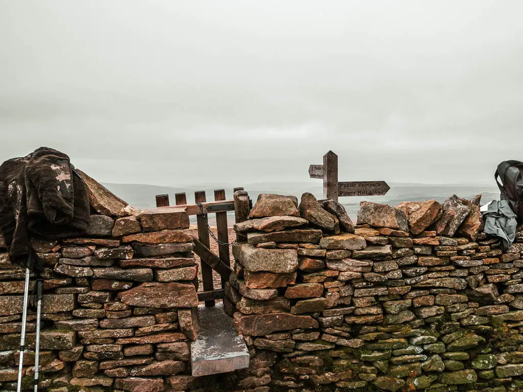 A stone wall with a small wooden gate across a small gap. There is hiking gear resting on the wall.