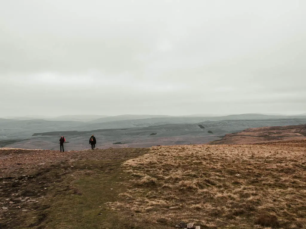 A wide grass trail with a couple of people walking along itched, and a view to the vast landscape of green hills way in the distance.