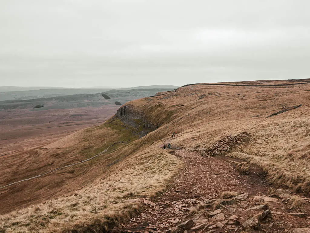 A dirt, rocky trail along the right with a view down the hill to the left, and to the cliffside ahead.