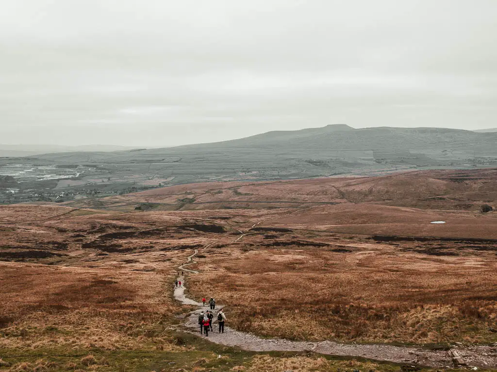 A long trail snaking downhill and ahead as far as the eye can see, across the undulating brown and orange landscape, on the walk down from Pen-y-Ghent. There are dark green hills way on the distance. There are lots of people walking along the trail.