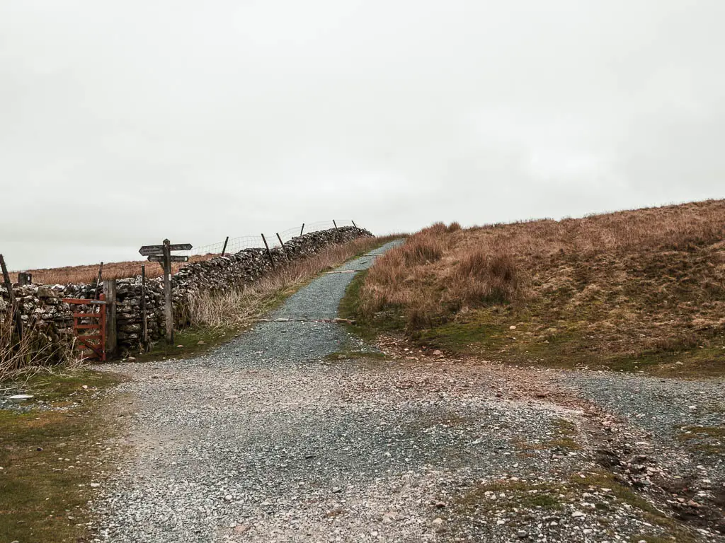 A gravel trail junction, with a stone wall on the left and a wooden trail signpost pointing in multiple directions next to it.