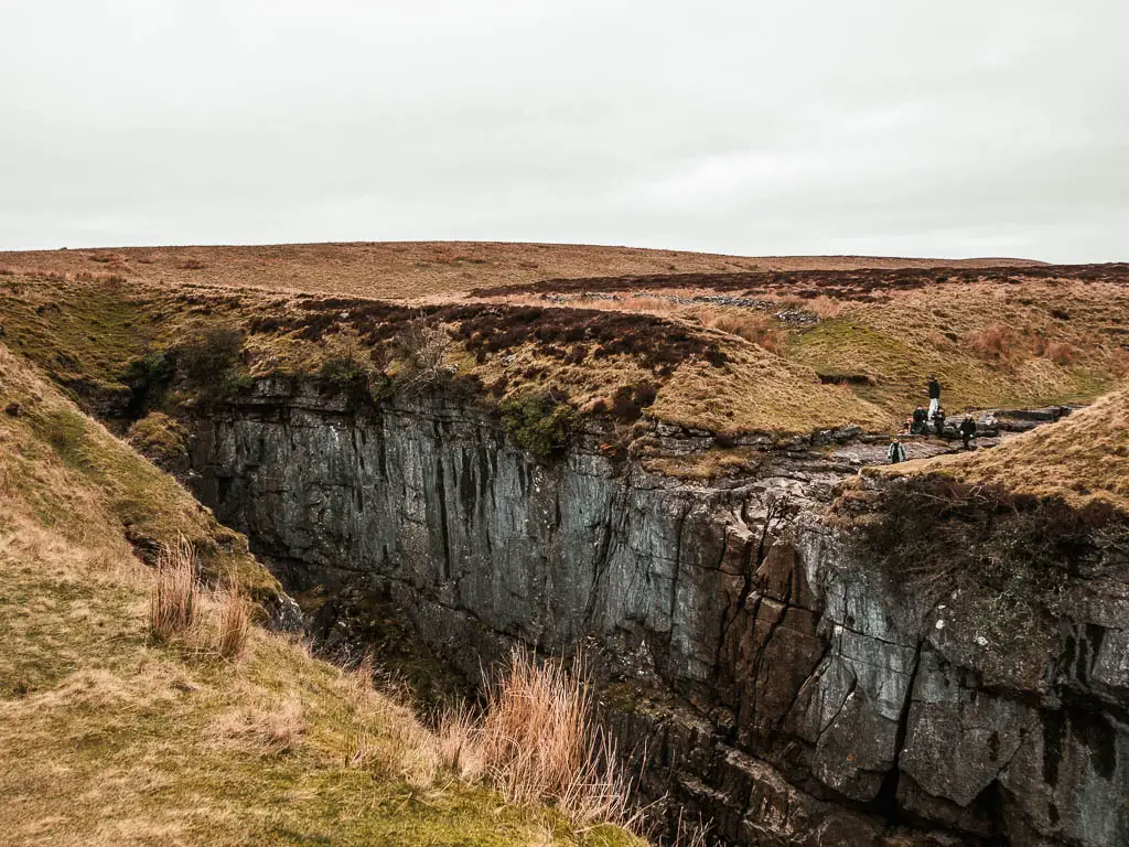 Looking down to the cliffside of the deep grass top covered cavern of Hull Pot. There are a few people on the other side.