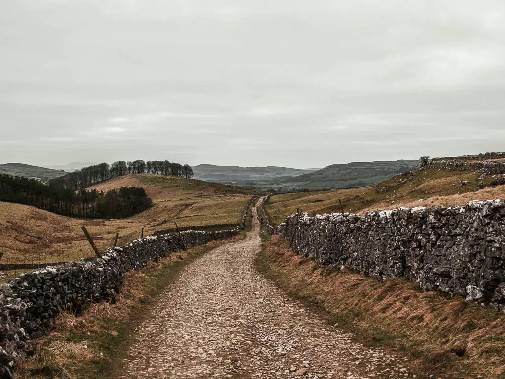 A long winding gravel stone trail, lined with stone walls, snaking across the grass hills ahead.