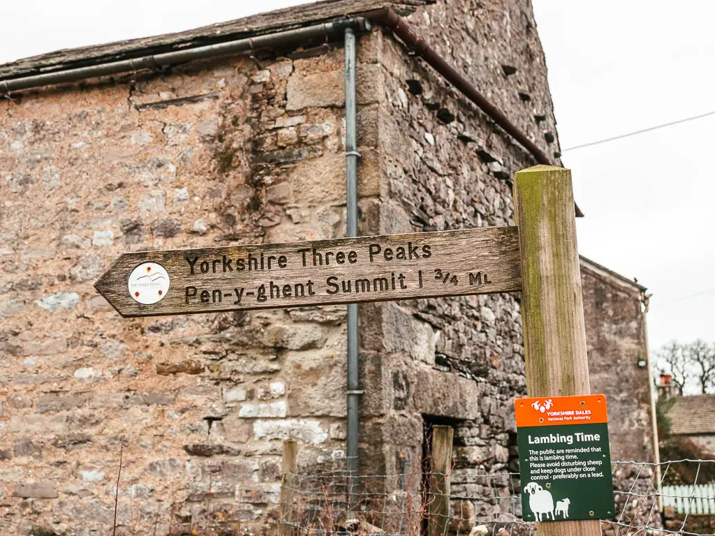 A wooden trail sign post in front of a stone building, which says ' Yorkshire three peaks, Pen-y-Ghent summit' at the start of the walk. 