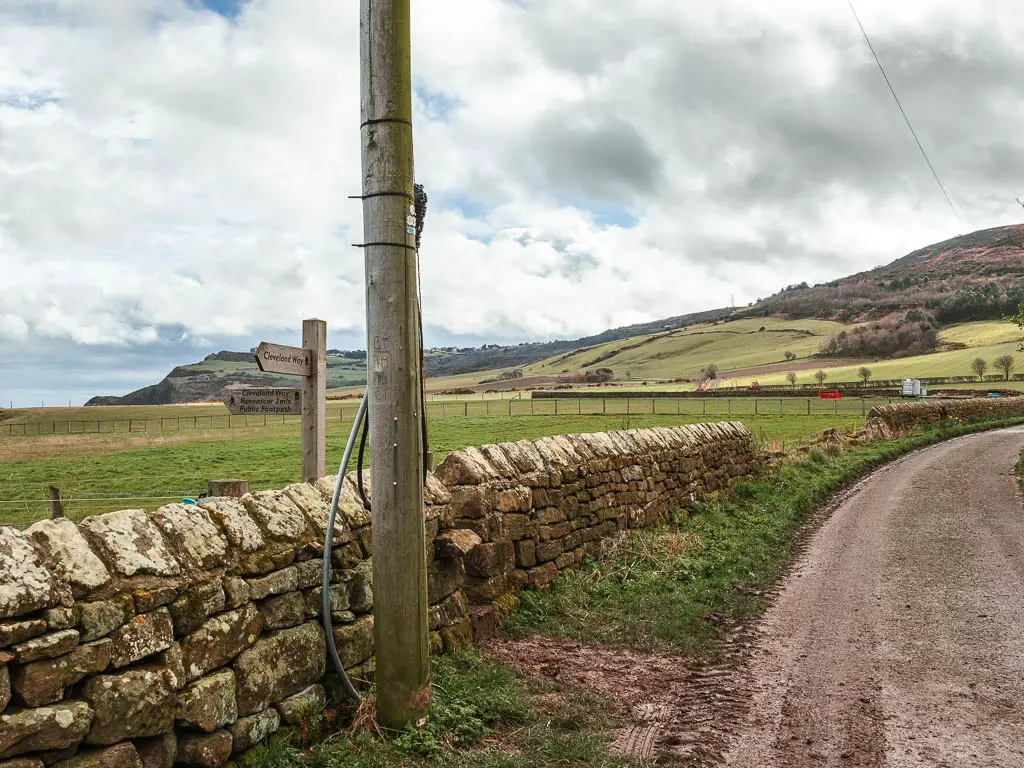 The road curving around to the right, and a stone wall on the left, with a wooden trail signpost on the other side of the wall.