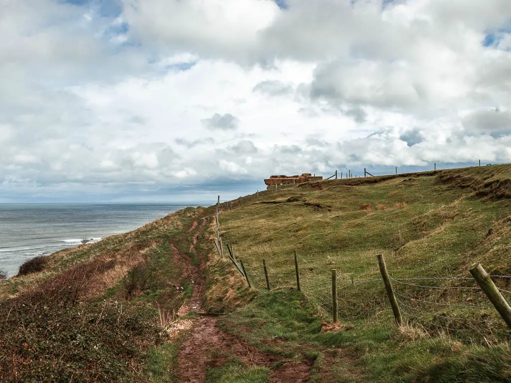 A rigged dirt trail on the side of a hill, with a wire fence to the right and the top of a bunker visible ahead.