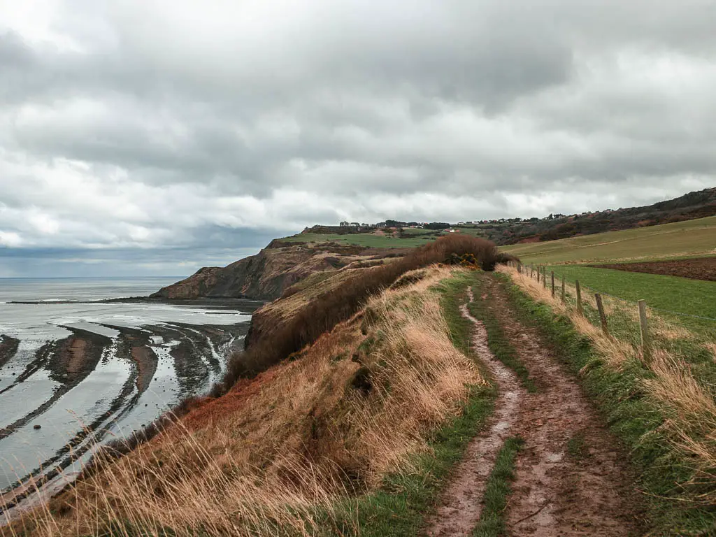 A muddy trail along the clifftop, with a view down to the sea to the left on the Robin Hood's Bay and Ravenscar walk. The sea is forming strips of water. There is a hill and cliffs ahead in the distance.