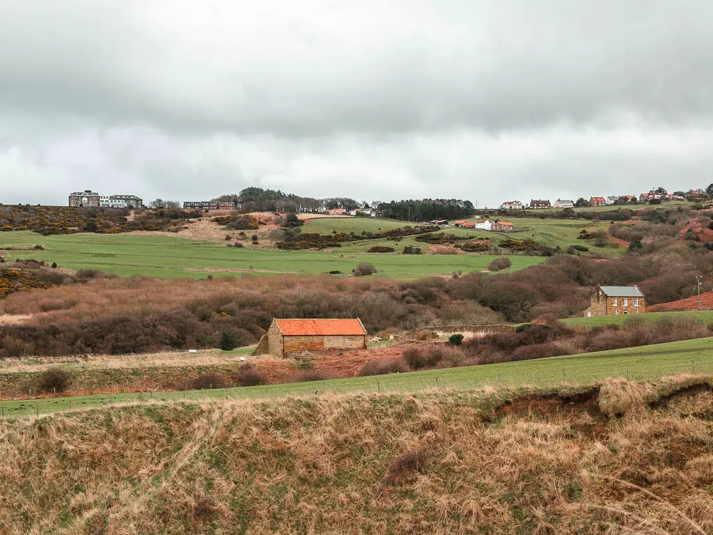 Looking across a field to a shed with an orange roof, and a hill beyond on. the circular Robin Hood's Bay and Ravenscar walk.