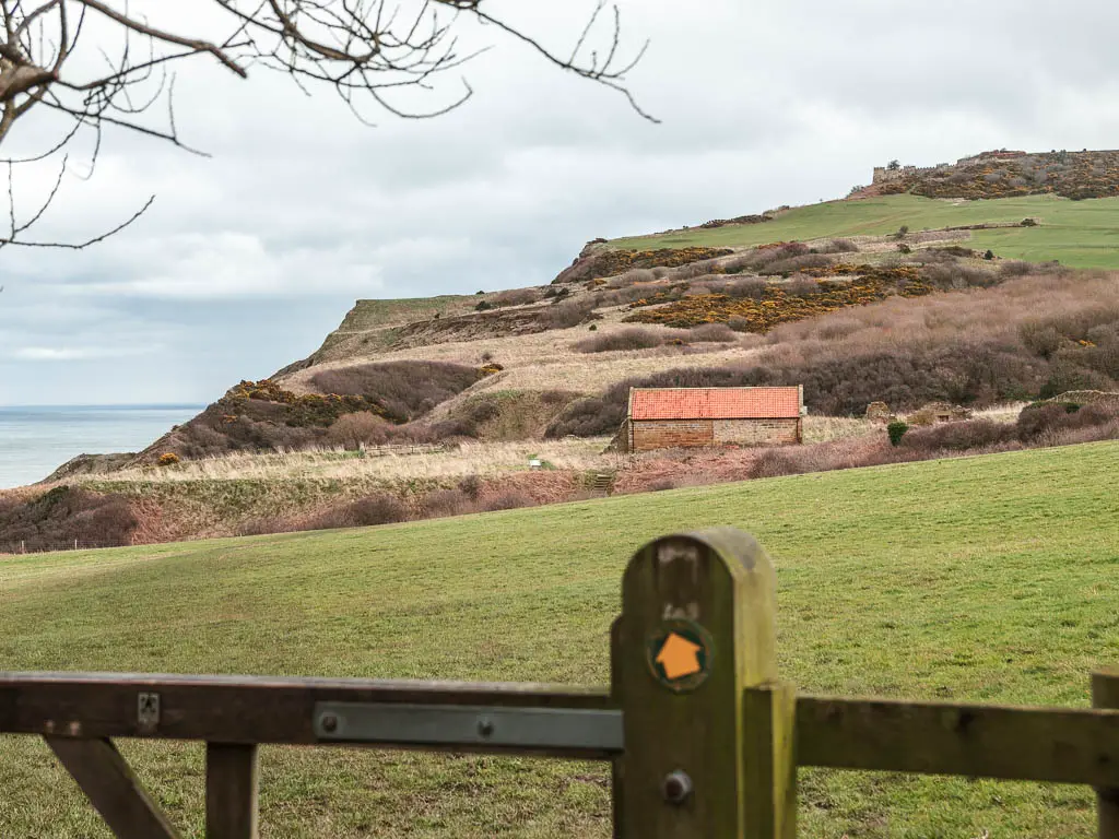 Looking over a fence with a yellow trail arrow pointing ahead to the field on the Robin Hood's Bay and Ravenscar walk. There is a hill on the other side of the field, and an orange roofed cottage shed.