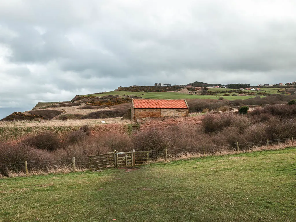 Looking across the field with bushes and a wooden gate in the wire fence on the other side on the walk towards Ravenscar from Robin Hood's Bay. There is an orange roofed shed past the fence.