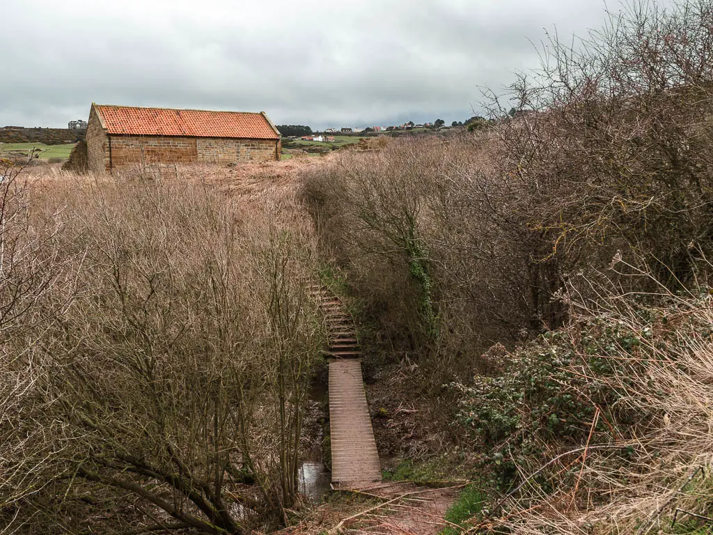 Looking down to a wooden plank walkway bridge and steps on the other side, leading up to an orange roofed stone walled shed cottage. The steps and wooden walkway are surround by leafless trees and bushes.