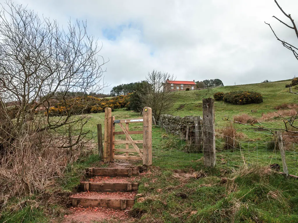 A wooden gate up a few steps, leading into a small grass field. There is a red roofed cottage at the top of the field.