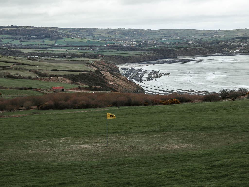 Looking down across the immaculately cut grass of the golf course, towards the cliffs and beach below. There is a yellow flag on a white pole in the middle of the golf course.