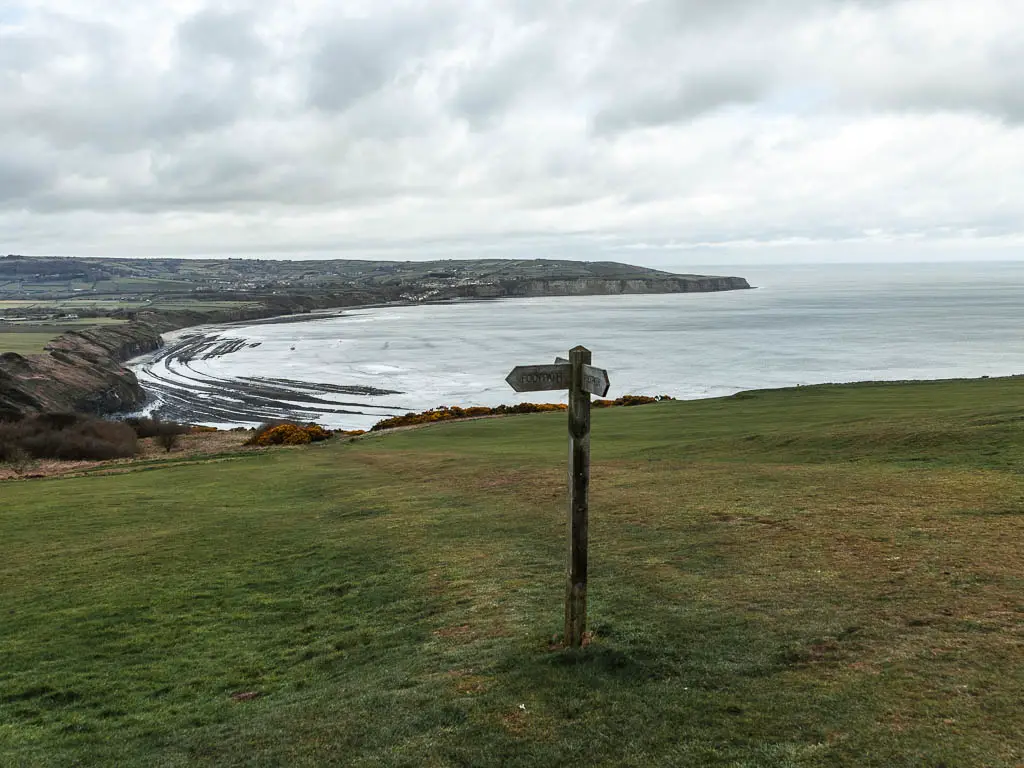 A wooden trail signpost pointing down a large grass field, with the sea in the distance. 