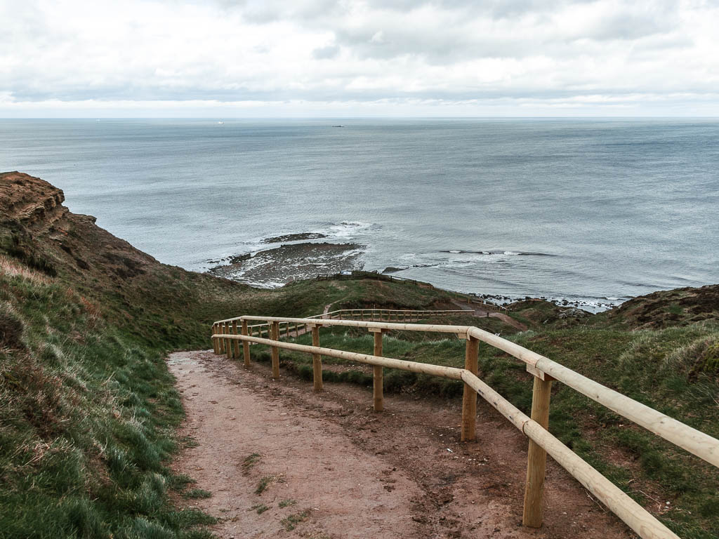 A dirt trail winding downhill, lined with a grass hill bank on the left and a wooden railing on there right, on the walk down to Ravenscar from Robin Hood's Bay. The sea is ahead at the bottom.