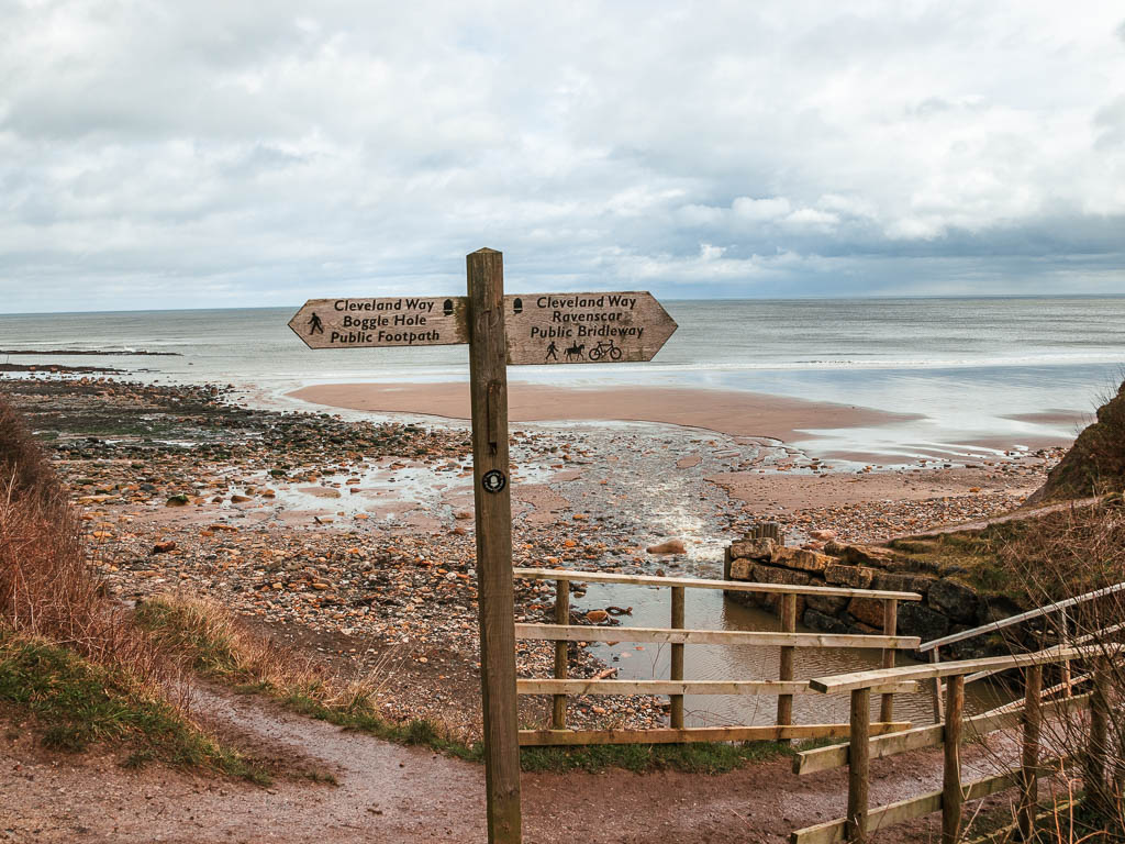 A wooden trail signpost marking the Cleveland way, with the sandy and rocky beach behind.