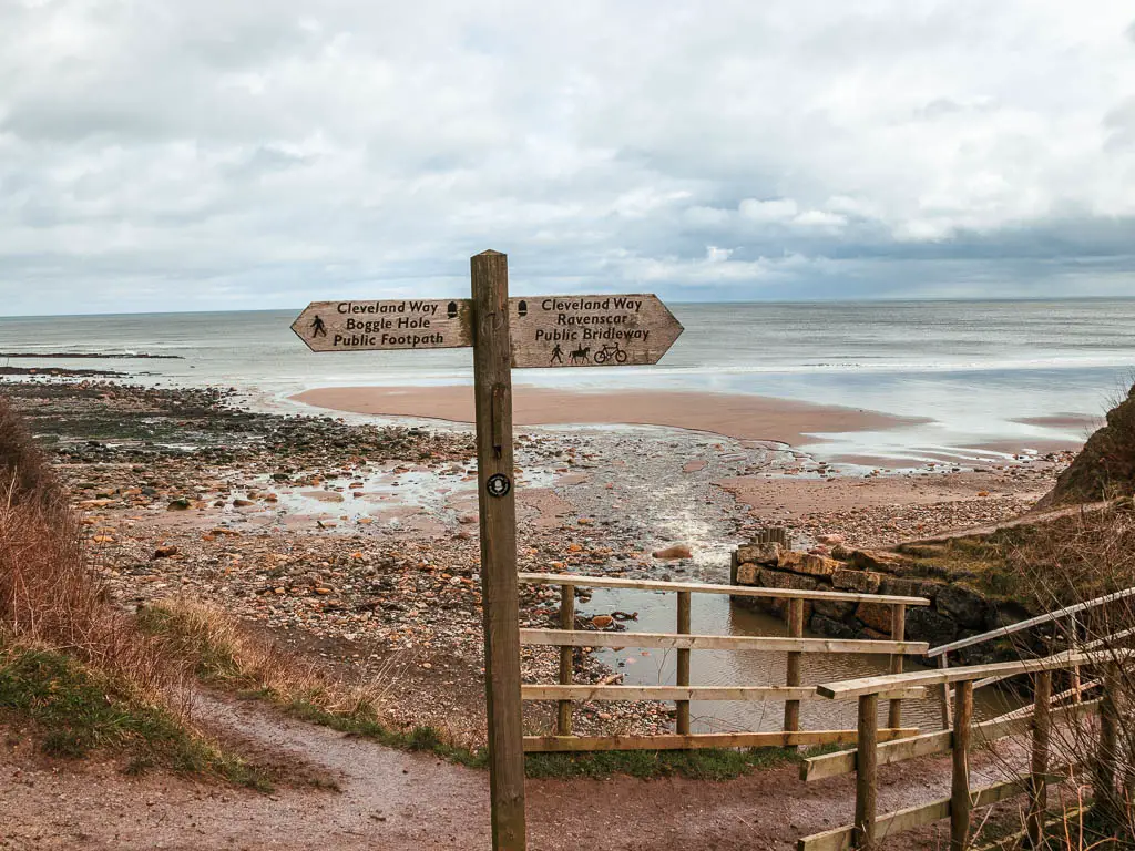 A wooden trail signpost marking the Cleveland way, with the sandy and rocky beach behind.