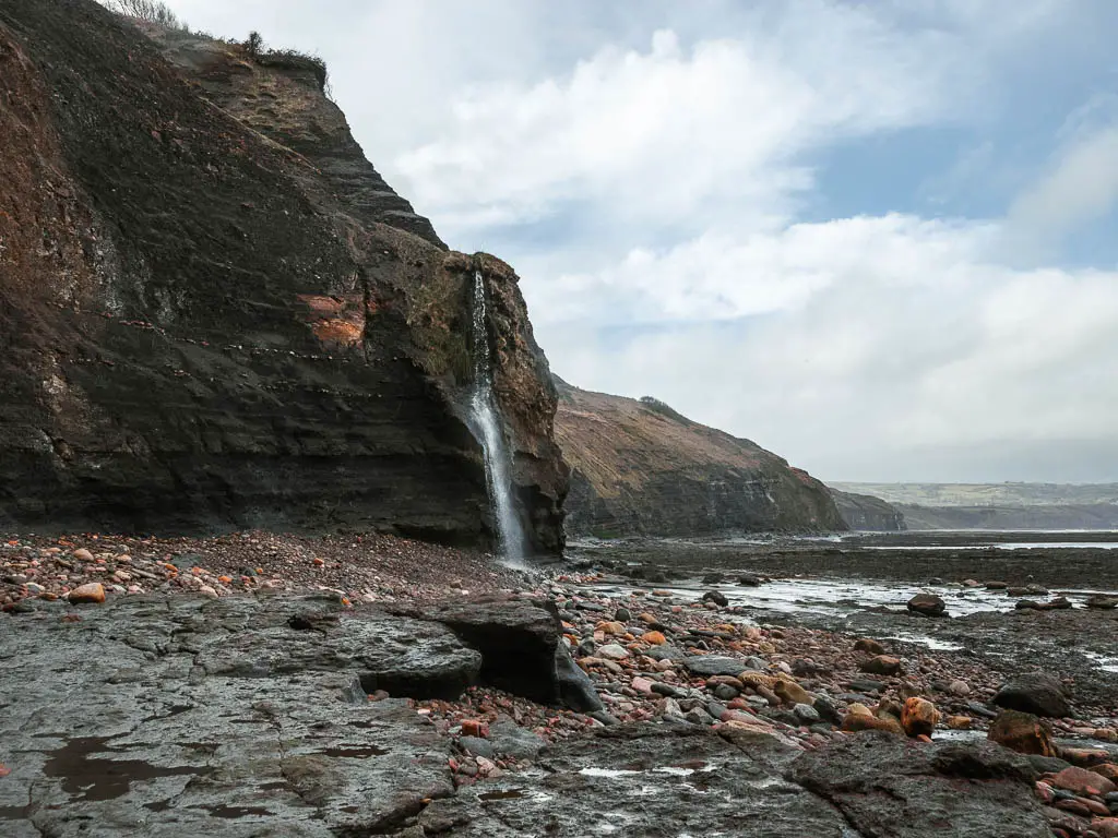A rocky beach below a tall cliff with a waterfall gushing down it, on the walk back to Robin Hood's Bay from Ravenscar. 