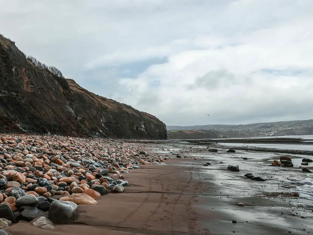 Looking along the sandy beach with large pebbles to the left below a dark cliff, on the walk towards Robin Hood's Bay from Ravenscar.