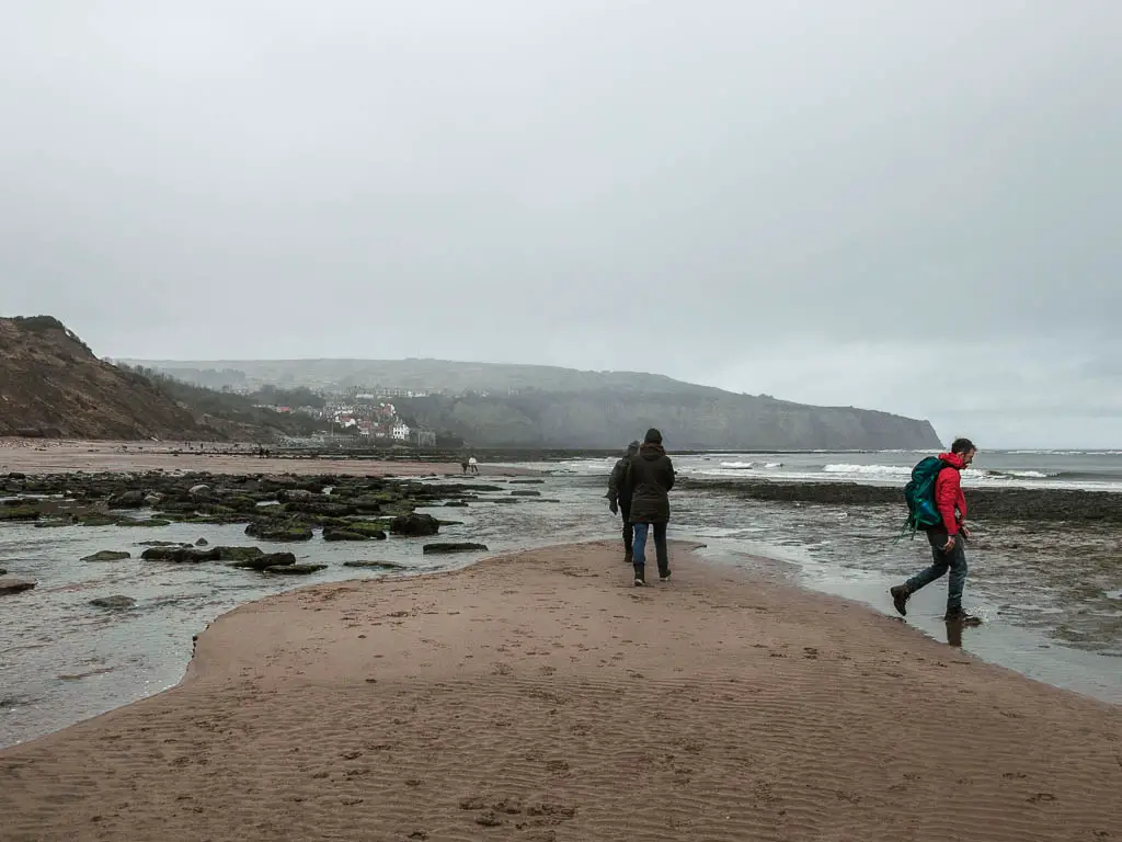 The sand surround by water and a view to the cliffs and Robin Hood's Bay in the distance. There are a few people walking on the sand pacts.