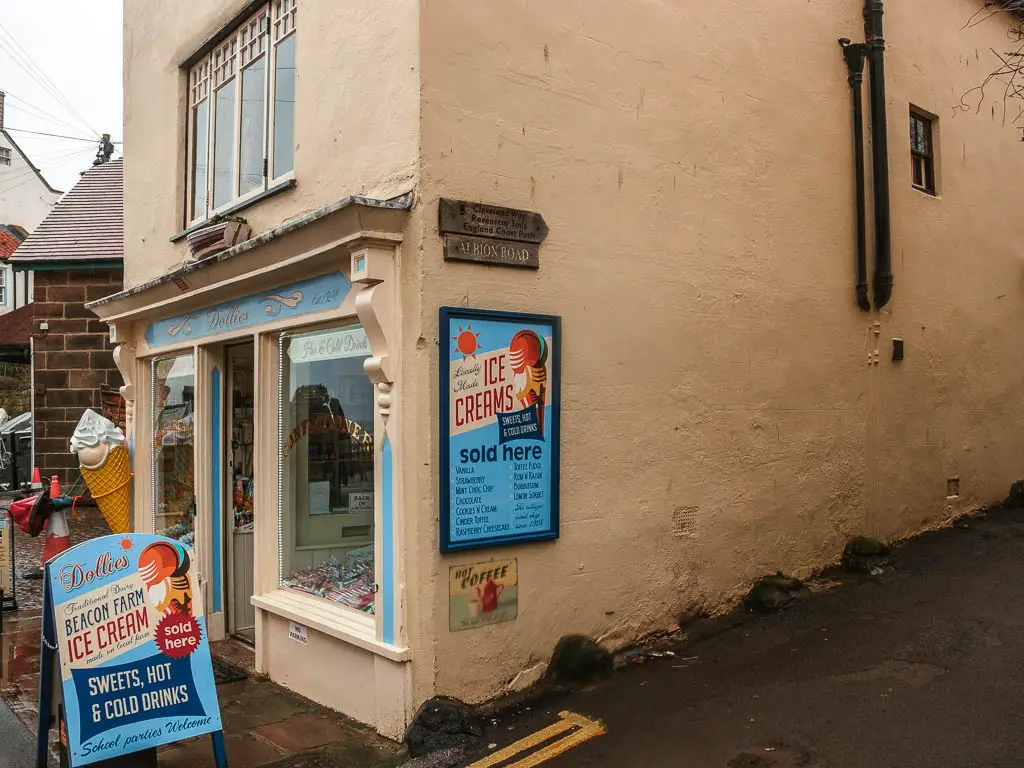 A yellow coloured corner ice cream shop, with blue ice cream shop signs outside. There is a wooden trail sign on the side of the wall.