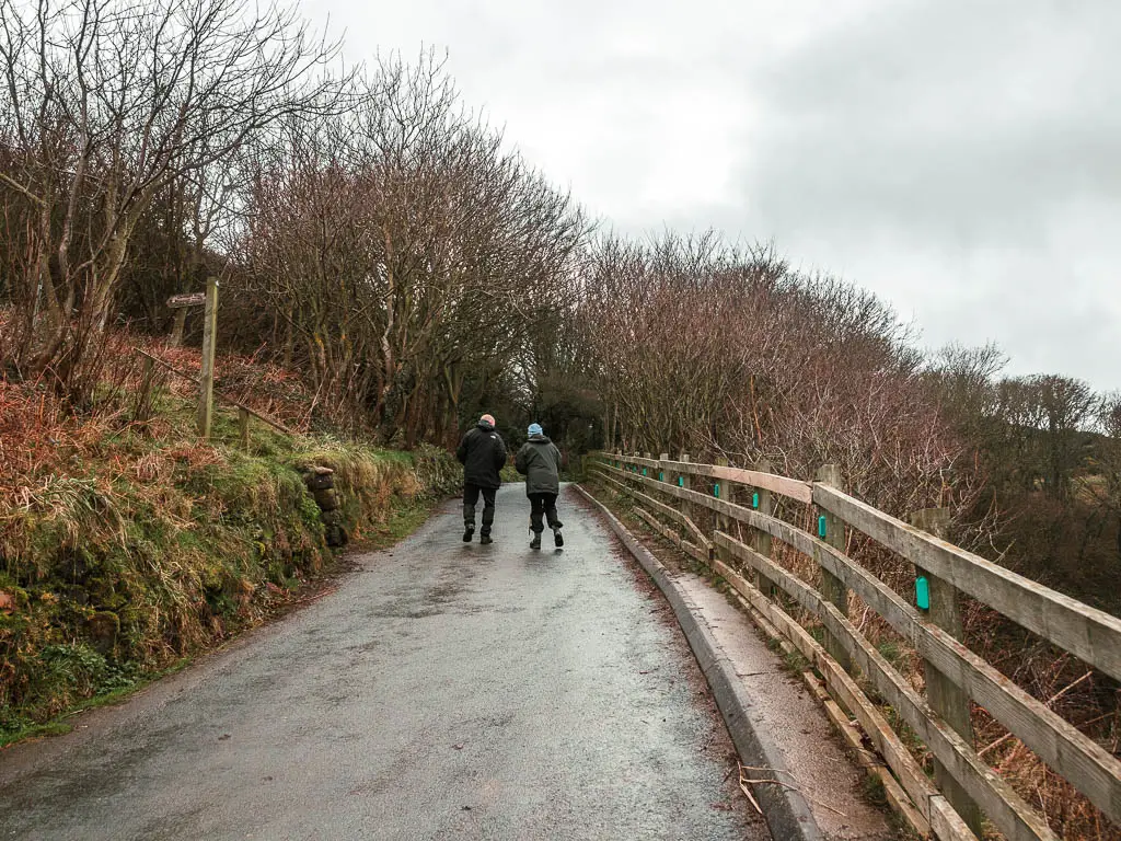 A road leading uphill, with a grass hill bank to the left and a wooden railing to the right. There are two people walking up the road. There is a wooden trail signpost pointing left up the bank.