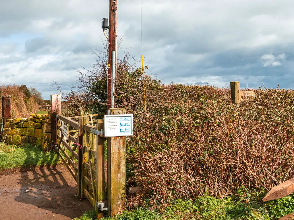 A wooden gate next to a bush. There is a wooden trail signpost poking up on the other side of the bush.