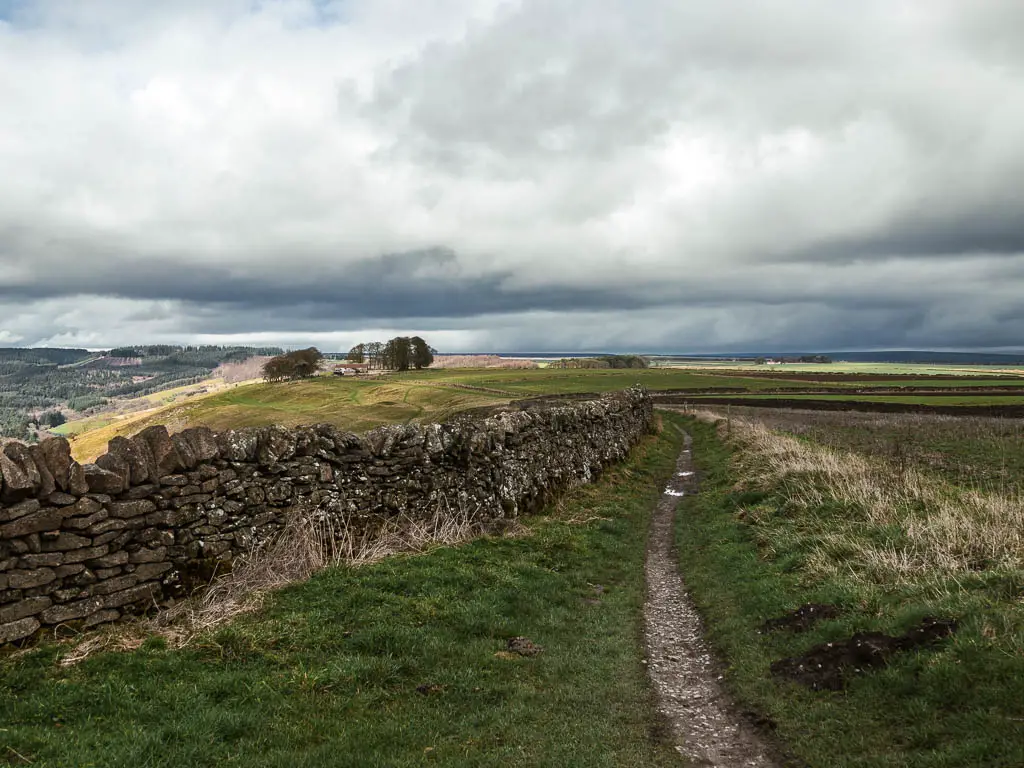 A narrow trail running along a stone wall to the left and field to the right, with a view to the hill side ahead.