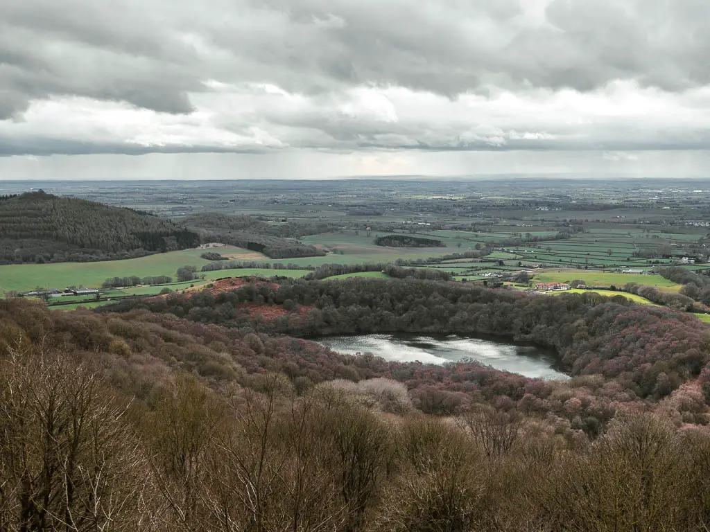 Looking down to Gormire lake surround by woodland trees, at the top of Sutton Bank. There is a pointy tree covered hill ahead to the left.