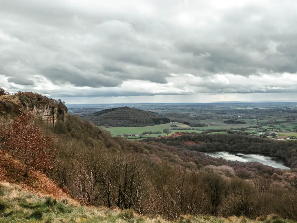 Looking down and towards the cliff side of Sutton Bank, and across the North York Moors ahead, halfway through the walk. Gormire lake is at the bottom surround by trees, and a pointy hill is in the distance.