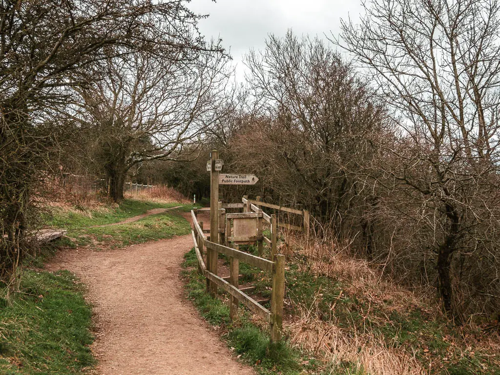 The trail to the left and wooden fence and wooden sign post on the right, pointing down the hill.