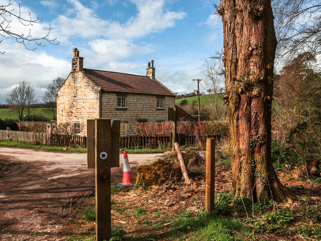 A road corner, with a brick house, surround by a wooden fence. The sky is blue with a few white clouds.