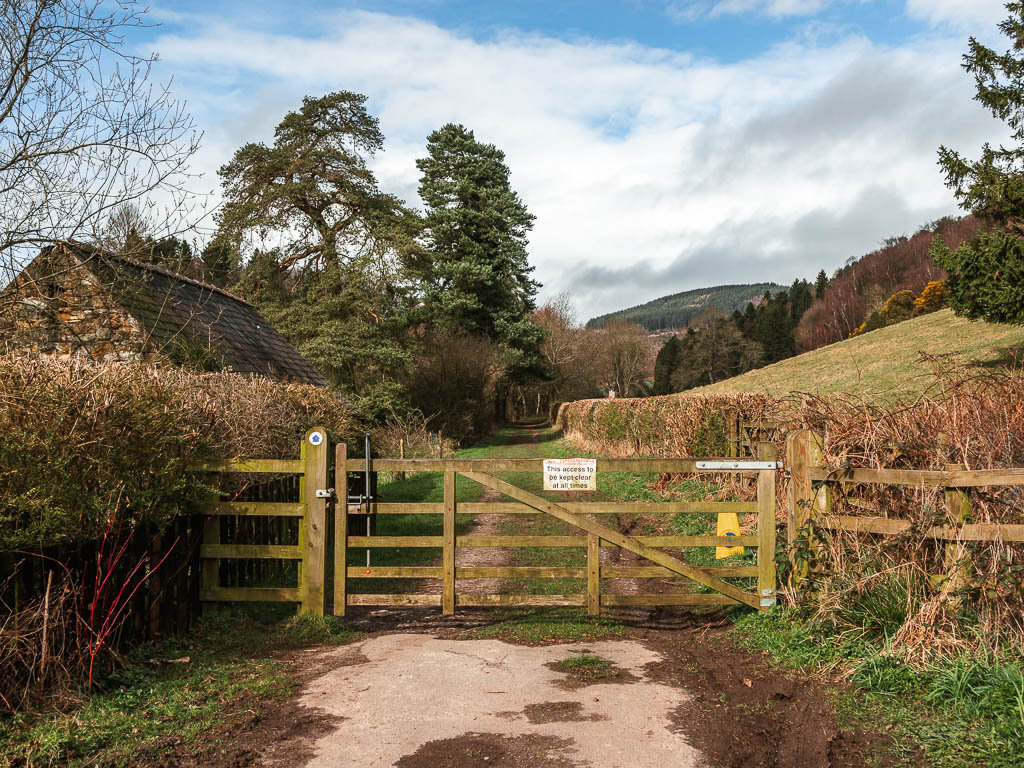 A wooden gate leading onto a grass type trail, with hedges lining the trail, and the rooftop of a houses to the left on the hedge.