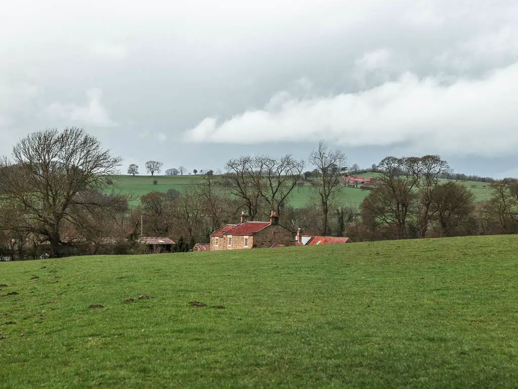 Looking across a grass field, with the top of a house with a red roof ahead, on the circular walk back from Sutton Bank.