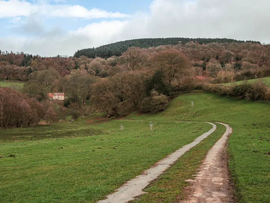 A track leading ahead and to the left, with a tree covered hill ahead, and a house partially hidden within the trees.