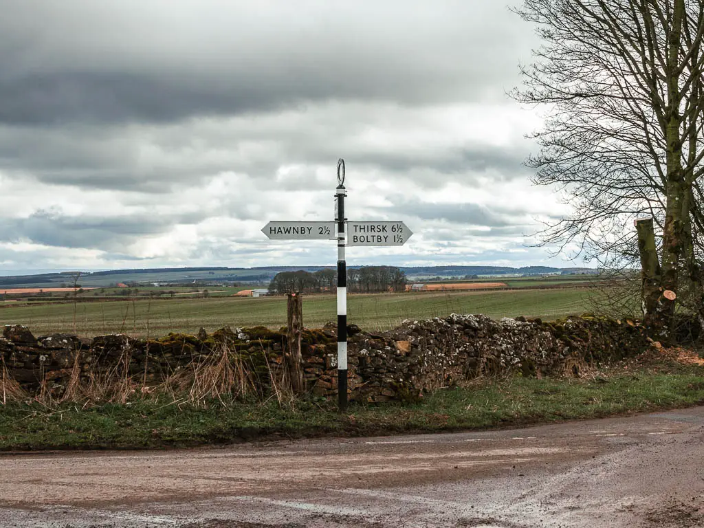 A road signpost with a black and white stripped pole, pointing left to Hawnby, and right to Boltby. The sign is on the side of a road, with a stone fence and field behind it.