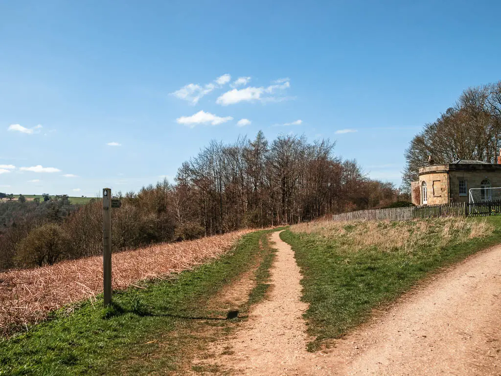 A dirt path leading straight with rough the grass, with a small outbuilding to the right, and a wooden trail signpost on the left side of the trail.