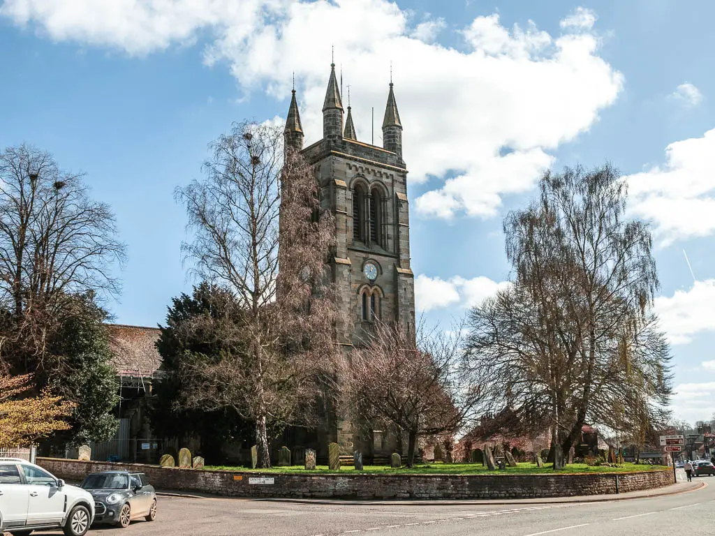 A church next to there road, with a graveyard on the green in front. The sky is blue with a few white clouds. There are some cars parked on the left.