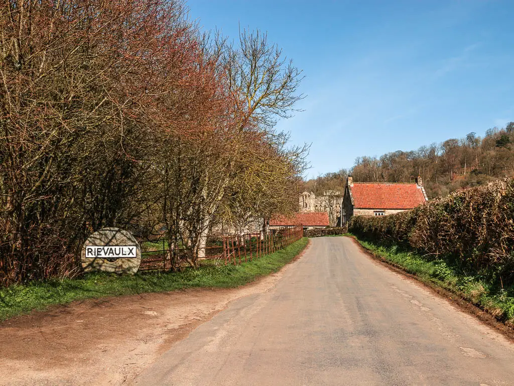 A country road leading straight, with some red roofed cottages ahead. There is a sign on the right saying Rievaulx. There is a hedge lining the right side of the road, and woodland on the left.