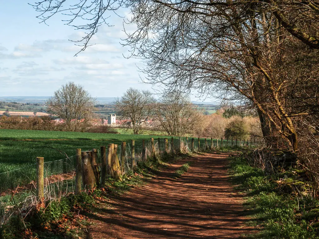 A dirt trail with woodland to the right, and a wire fence and field to the left with a view down to Helmsley on the walk back from Rievaulx Abbey.