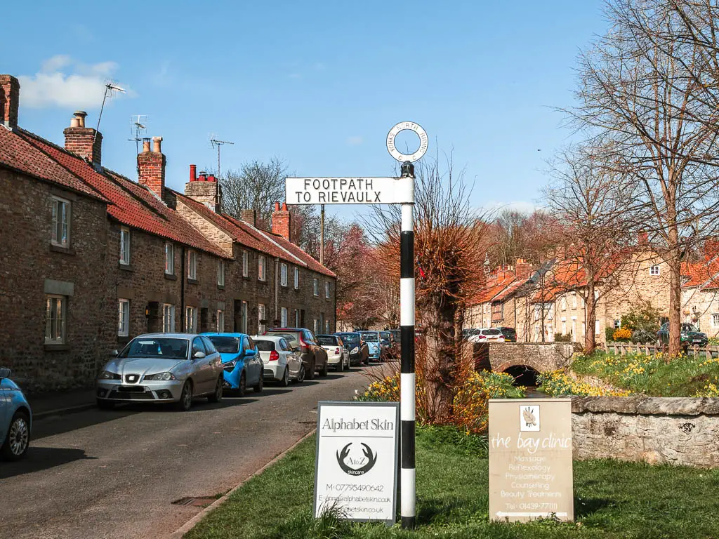 A black and white stripped pole with a white sign saying 'Footpath to Rievaulx', at the start of the walk from Helmsley. The sign is on a green, with a road and row of houses to the left. There are cars parked all along the road side.