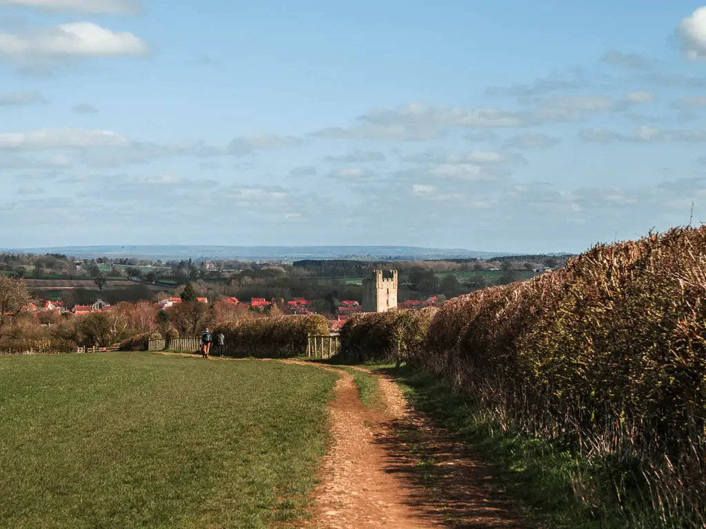 A dirt trail along the right side of a grass field, with a hedge to the right of it, and a view down to the village of Helmsley ahead. 