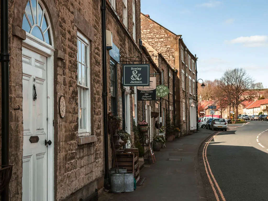 A road on the right, and stone walled shops and store fronts on the left in Helmsley. 