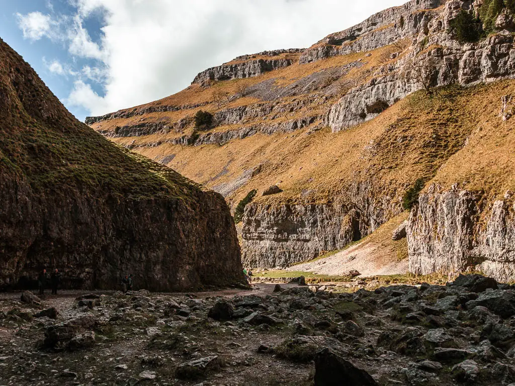 Looking through the gorge of Gordale Scar, across a ground covered in rocks. The sun is shining down on the cliffside outside the gorge.