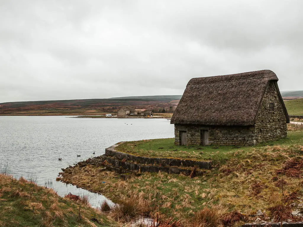 A thatched roofed barn on the green, on the edge of the reservoir.