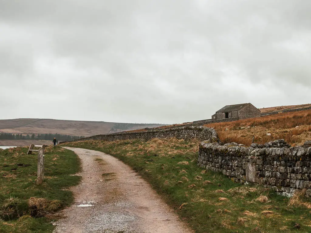 A wide path curving ahead to the left, lined with grass banks, and a stone wall on the right. There is a barn up the hill to the right.