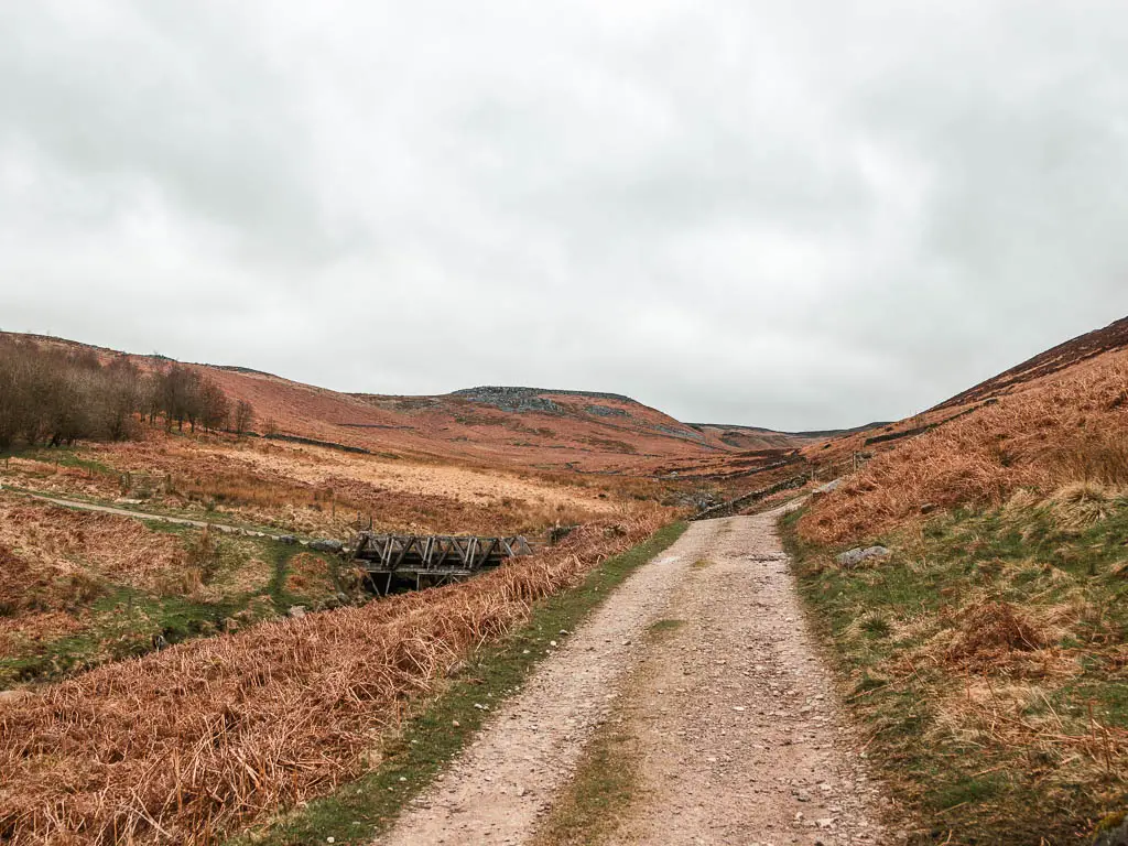 A gravel type path leading through a dip in the hills, and a small wooden bridge ahead to the left.