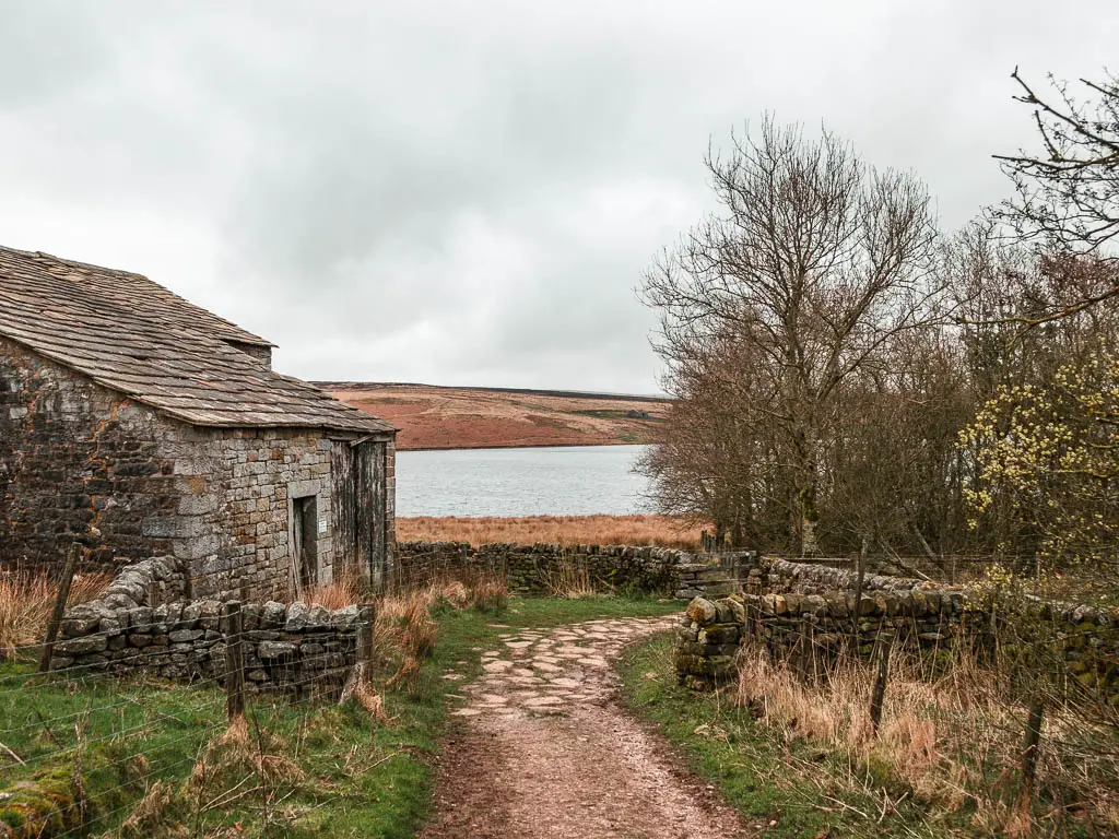 The path leading down and past a stone walled barn, with the water visible through a gap between the barn and trees.